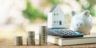 Stack of coins, piggy bank and calculator in front of model house. How much do house buying companies really pay for your home?